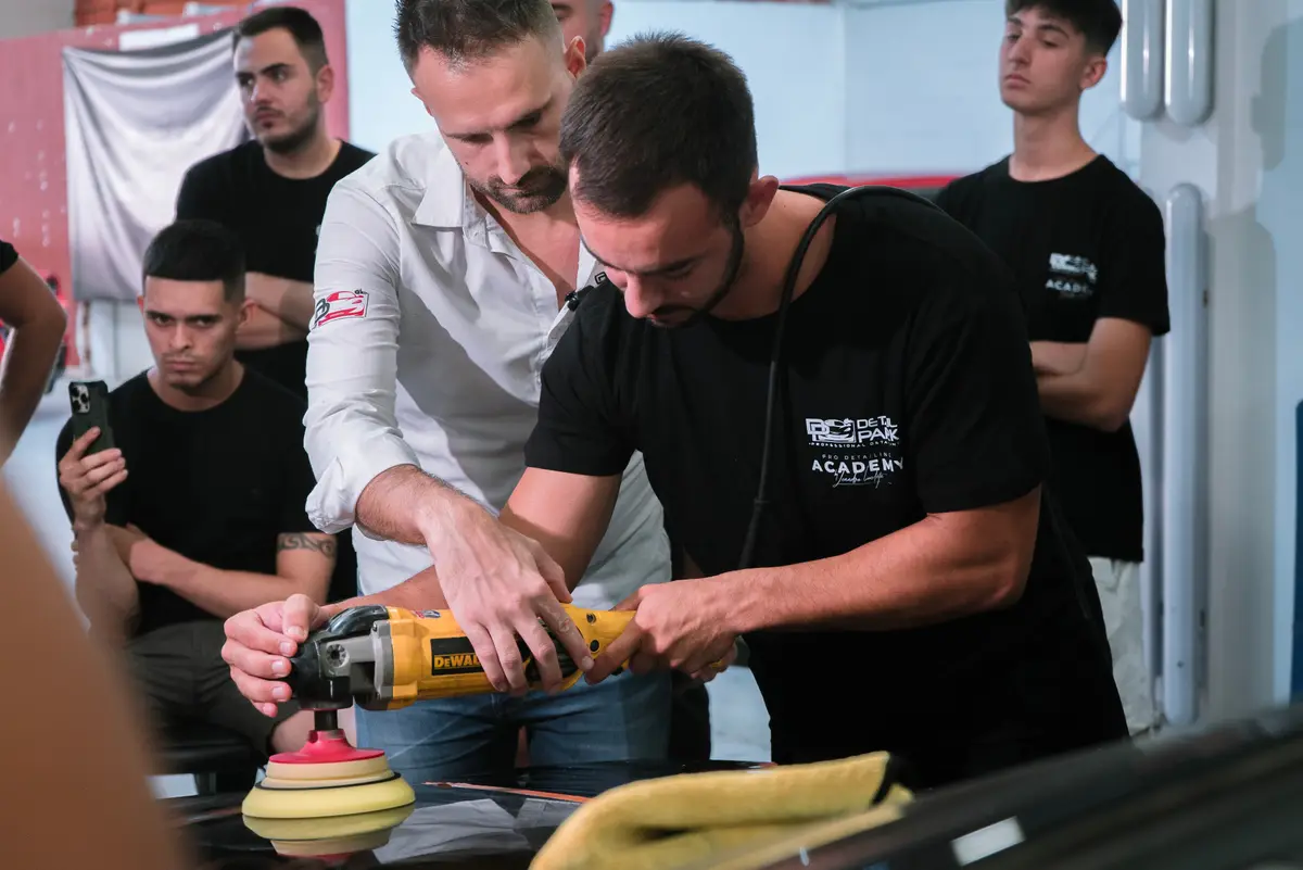 Alumno puliendo un coche con una pulidora DeWalt mientras el formador le guía y otros alumnos graban con el móvil