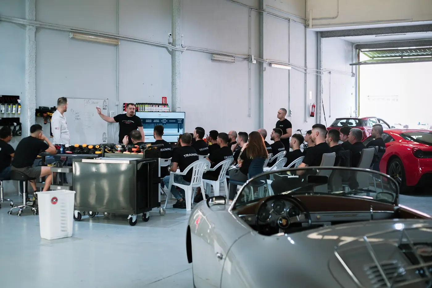 Clase teórica con pizarra y pantalla junto a un Porsche clásico en el taller